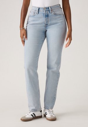Personne portant un jean droit bleu clair et des baskets blanches avec des rayures noires, se tenant devant un fond clair.