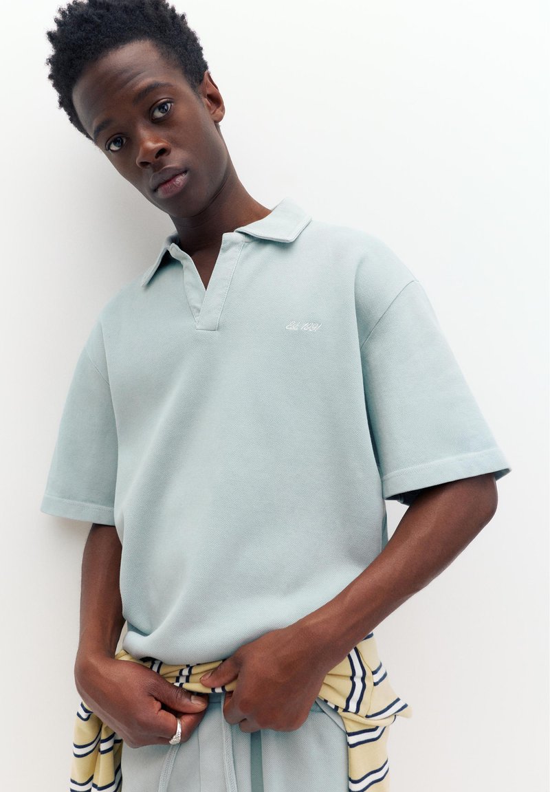 Jeune homme aux cheveux bouclés et foncés porte un polo bleu clair à manches courtes et tient un pull rayé jaune et bleu marine autour de sa taille.