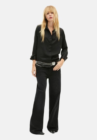 Schwarze Satinbluse mit durchgehender Knopfleiste, kombiniert mit hoch taillierten, dunkel ausgeweiteten Hosen mit einem silbernen Kettengürtel-Detail.