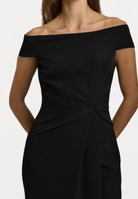 Černé šaty s odhalenými rameny, přiléhavým korzetem, draperií a detailním uzlem v pase. Hladká textura, délka po kolena.