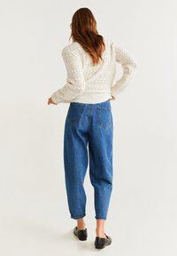 Femme aux cheveux longs portant un pull blanc en maille, un jean taille haute bleu et des ballerines noires, debout, les mains sur les hanches, vue de dos.