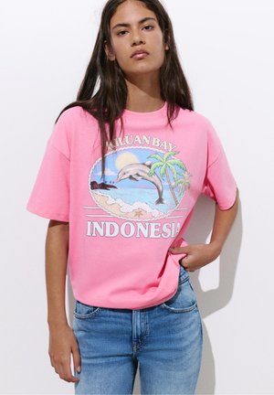 Fiatal nő rózsaszín pólóban, melyen delfin, pálmafák, tengerparti jelenet és „KILUAN BAY INDONESIA” felirat látható, kék farmerrel párosítva.