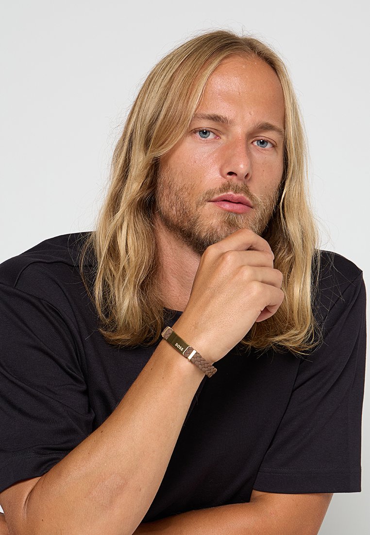 Bracelet en cuir tressé brun avec un fermoir en métal doré portant l'inscription "BOSS" gravée. Porté au poignet, présenté sur un fond simple.