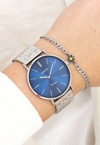 Montre en argent avec un cadran rond bleu, bracelet en maille argentée élégant, marqueurs horaires minimalistes et le nom de la marque en dessous de la position de 12 heures.