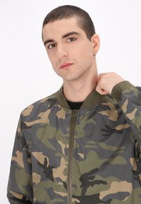 Camo bomberjakke i grønne, beige og sorte nuancer. Har lynlås lukning, ribbede manchetter og design med rund hals. Glat stofstruktur.
