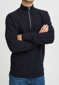Pulover cu fermoar bleumarin, cu guler înalt, model tricotat texturat și manșete și tiv cu nervuri, purtat cu blugi albaștri.