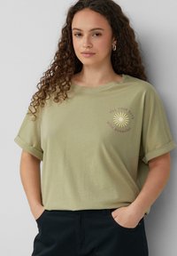 Olijfgroene katoenen T-shirt met opgerolde mouwen, voorzien van een bedrukt zonnenmotief en de tekst "VUL JE DAGEN MET ZONNESCHIJN" in beige.