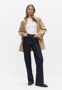 Beige regenjas over een wit T-shirt, donkerblauwe uitlopende jeans met een bruine riem en bruine schoenen. Soepele stoftextuur en een getailleerde pasvorm.