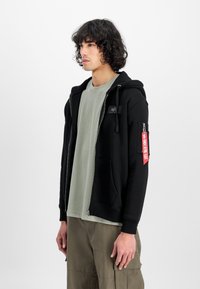 Sort zip-up hættetrøje med en tekstureret finish, prydet med et logo patch, sidelommer og et rødt mærke på venstre ærme. Bæres over en lys grøn skjorte.