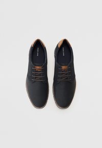 Zapatos negros con tela texturizada, acentos de cuero marrón y cordones marrones. Este diseño presenta una punta redondeada y un detalle de costura en contraste.