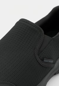 Black slip-on shoes made of textured mesh material, featuring a rounded toe and a subtle Skechers logo on the side.