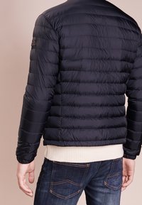 Tmavě modrá péřová bunda s žebrovaným krémovým lemem a manžetami, s vodorovnými prošívanými panely. Nositelná přes tmavé denimové džíny.