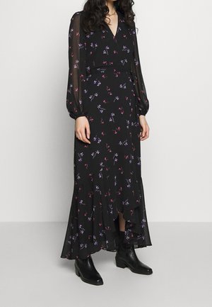 Frau in einem langen schwarzen Blumenkleid mit transparenten langen Ärmeln und schwarzen Stiefeletten, steht vor einem schlichten hellen Hintergrund.