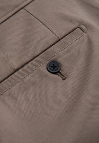 Beige katoenen stof met een gestructureerde afwerking, met een close-up van een donkere knop en een gestikt pocketdetail.