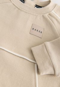 Beige sweatshirt med rund halsudskæring, der har et lille "BAKER" mærke, hvide piping langs sømmene og tekstureret stof.
