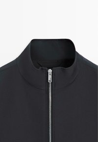 Schwarze Jacke mit Stehkragen und silbernem vorderem Reißverschluss, dargestellt vom Hals bis zur Mitte der Brust vor weißem Hintergrund.