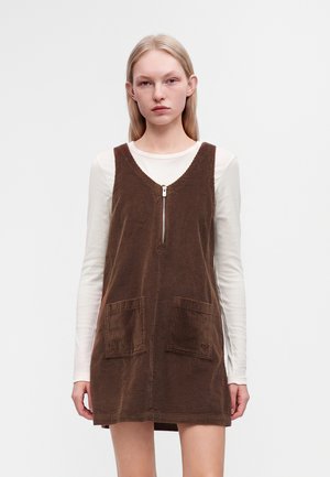 Jeune femme aux longs cheveux blonds portant une robe marron en velours côtelé sans manches par-dessus une chemise blanche à manches longues, debout devant un fond uni.