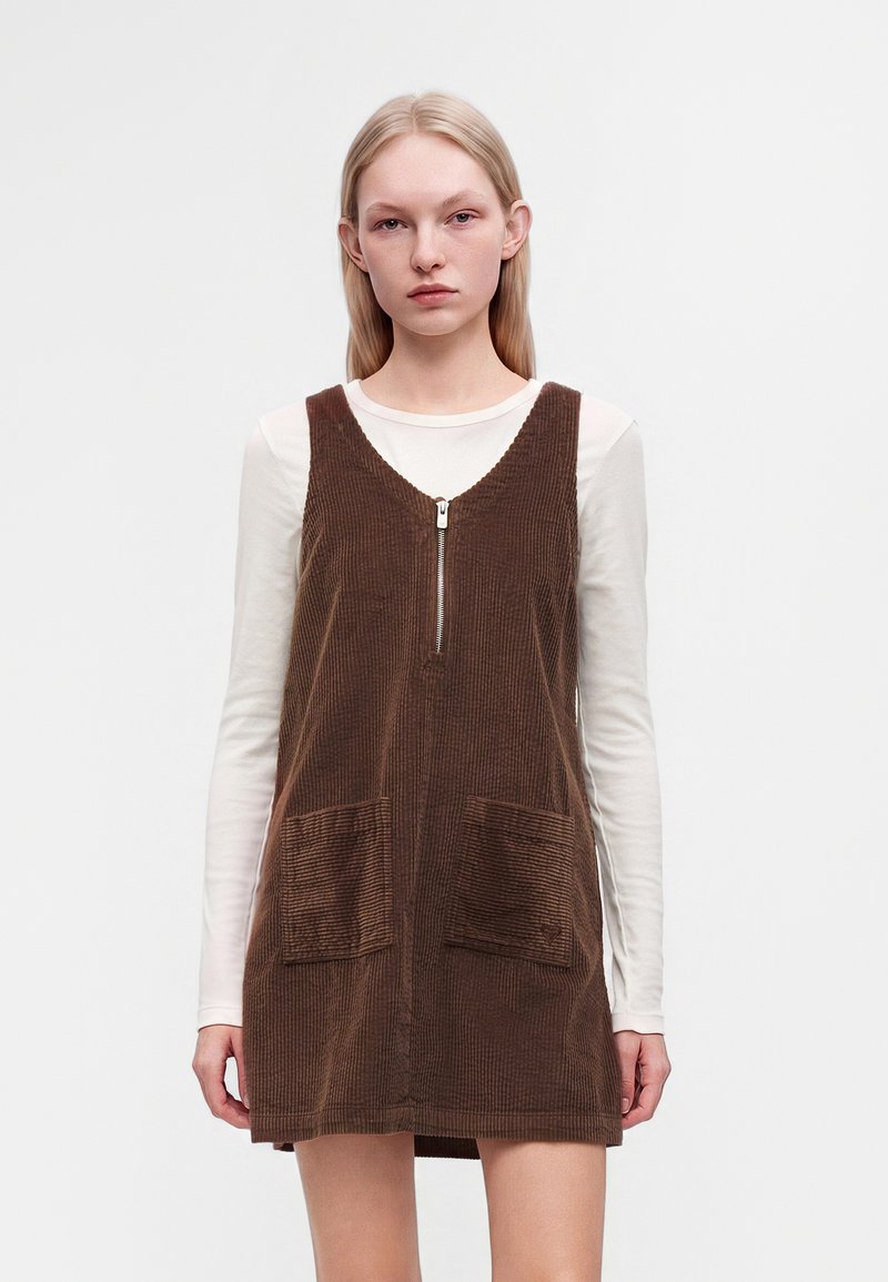 Jeune femme aux longs cheveux blonds portant une robe marron en velours côtelé sans manches par-dessus une chemise blanche à manches longues, debout devant un fond uni.