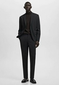 Wysoki mężczyzna w czarnym garniturze i czarnym golfem, stojący z jedną ręką w kieszeni na jednolitym szarym tle.