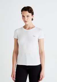 Tânără cu păr scurt și bronz, îmbrăcată într-un tricou alb mulat cu mânecă scurtă, având un mic logo negru pe piept și pantaloni negri.