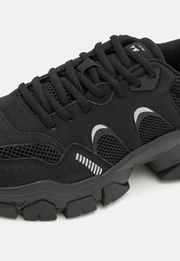 Chaussure de sport noire avec une tige texturée, des panneaux en mesh et des accents réfléchissants. Dotée d'une semelle épaisse et robuste ainsi que de lacets plats pour un ajustement sécurisé.