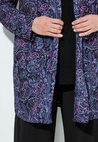 Cardigan lungo a fantasia in navy, con motivi paisley rosa e blu. Tessuto leggero con taglio dritto, indossato sopra una maglietta nera e pantaloni.