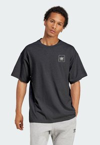 Tmavě šedé bavlněné tričko s volným střihem, krátkými rukávy a čtvercovou černobílou nášivkou s logem na levé straně hrudi.