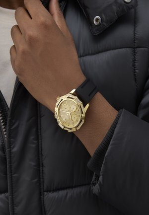 Orologio d'oro con cinturino in gomma nera, cassa ottagonale e quadrante d'oro testurizzato con tre sub-quadranti e dettagli in cristallo.