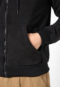 Schwarzer Fleece-Hoodie mit durchgehendem Reißverschluss, zwei Fronttaschen sowie gerippten Bündchen und Saum. Weiche Textur mit glattem Abschluss am Reißverschluss.