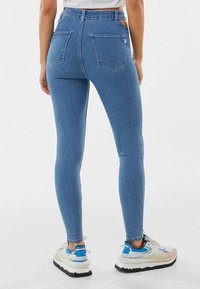 Jeans de mezclilla azul de cintura alta con textura suave, dos bolsillos traseros y costuras contrastantes, combinados con zapatillas blancas y coloridas.