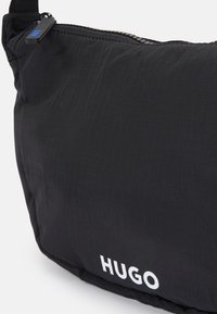 Borsa a tracolla nera realizzata in tessuto texture. Presenta una chiusura con zip e un prominente logo "HUGO" bianco sul fronte.