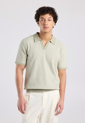 Jeune homme aux cheveux bouclés portant un polo en maille à manches courtes vert clair et un pantalon écru, sur un fond uni.