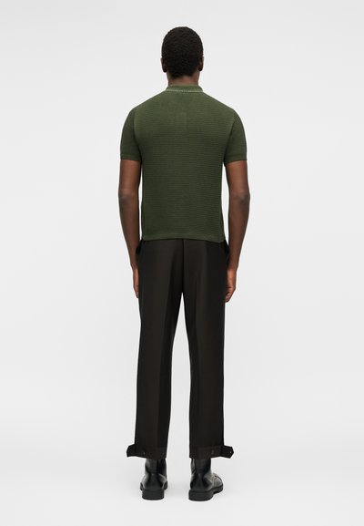 Άνδρας που φοράει μια κοντομάνικη πλεκτή μπλούζα ελιάς, μαύρο παντελόνι ραμμένο στα μέτρα με λουριά στα μανσέτα και μαύρες δερμάτινες μπότες, στέκεται γυρισμένος με την πλάτη.