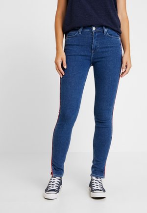 Femme portant un jean skinny bleu foncé avec des bandes latérales rouges, des baskets bleu marine avec des lacets blancs et un haut sans manches bleu foncé.