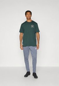 Žalsvi medvilniniai marškinėliai su mažu smėlio spalvos logotipu ant krūtinės, derinami su šviesiai pilkomis siaurėjančiomis sportinėmis kelnėmis su baltais dryžiais ir adidas logotipu.