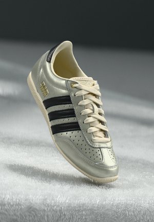 Light gray leather sneaker with black stripes, perforated details, beige laces, and a gold logo patch. Textured rubber sole.