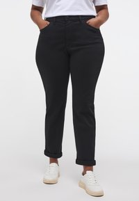 Fekete farmernadrág klasszikus szabással, gombos záródással, elülső zsebekkel és felhajtott szegéllyel, fehér sportcipővel párosítva.