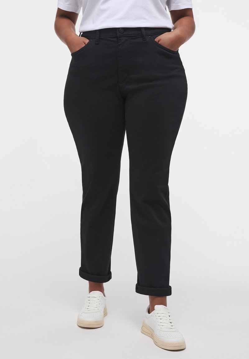 Fekete farmernadrág klasszikus szabással, gombos záródással, elülső zsebekkel és felhajtott szegéllyel, fehér sportcipővel párosítva.