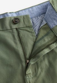 Pantalons vert olive avec une fermeture éclair à l'avant et un bouton métallique. L'intérieur est en tissu bleu clair avec des rayures blanches. Texture lisse.