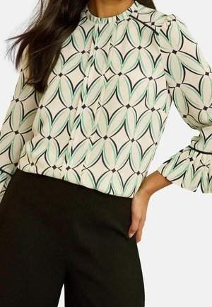 Donna che indossa una camicetta a maniche lunghe con motivo geometrico a foglie verdi e nere e pantaloni neri, una mano sui fianchi.