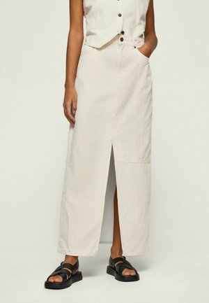 Femme portant une jupe longue couleur crème taille haute avec une fente à l'avant, associée à des sandales noires à bout ouvert et un gilet sans manches boutonné.