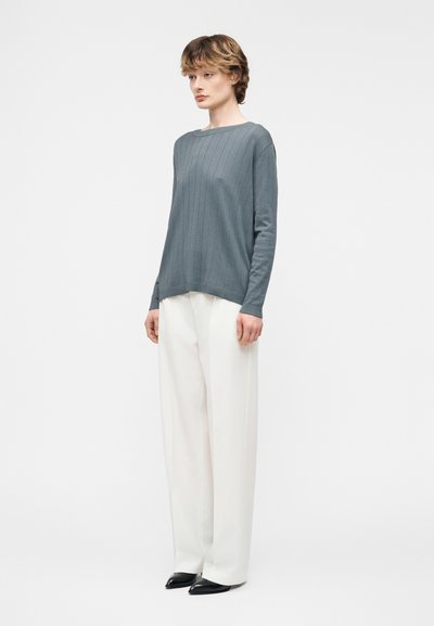 Νεαρό άτομο με κοντά μαλλιά που φοράει μπλε-γκρι ριγέ πουλόβερ, φαρδιά λευκά παντελόνια και μαύρα μυτερά παπούτσια, στέκεται μπροστά σε λευκό φόντο.