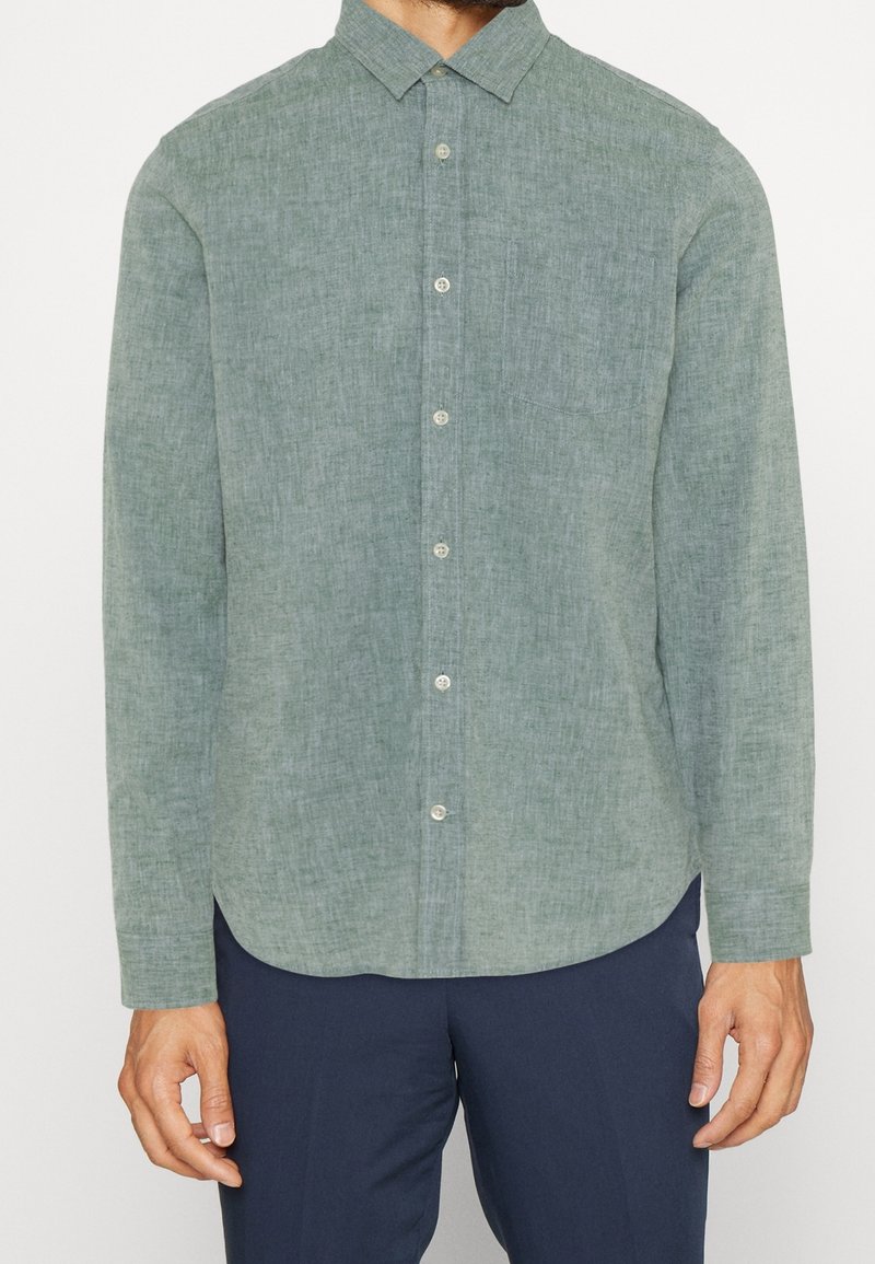 Homme portant une chemise en lin vert clair à manches longues, boutonnée, avec col et un pantalon bleu marine, debout devant un fond uni.