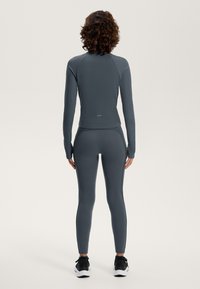 Temno siva športna majica in legice z rebrasto teksturo. Prilegajoča zasnova, dolgi rokavi in režami za palce. Oblečeno z črnimi supergami.