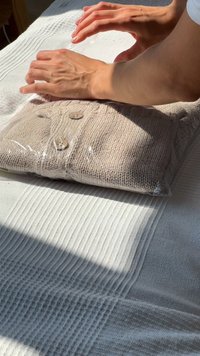 Beige neulepaita puuhelmillä, käärittynä läpinäkyvään muoviin, lepää vaaleanharmaalla teksturoituneella kangaspinnalla. Kädet avaavat sitä.