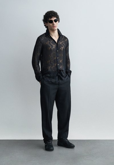 Jeune homme portant une chemise noire transparente à motifs, un pantalon noir, des baskets noires et des lunettes de soleil, debout les mains dans les poches.