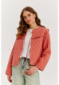 Kurtka pikowana w kolorze koralowym z okrągłym kołnierzem, luźnym krojem i detalami ze sznurkiem. Posiada falbany i długie rękawy. W połączeniu z jasnym dżinsem.