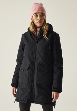 Mujer con un abrigo largo de invierno negro con capucha, gorro de punto rosa y suéter rosa con cremallera, mirando ligeramente hacia un lado sobre un fondo liso.