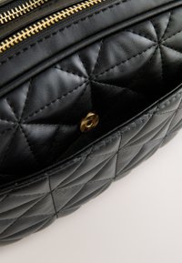 Sort quiltet lædertaske med et tekstureret mønster, guldfarvet lynlås og en trykknapdetalje foran.