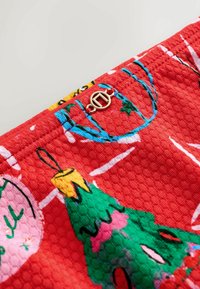 Piros textúrájú szövet karácsonyfás mintával zöld és rózsaszín színekben, fehér kiegészítőkkel és arany kör alakú hardver részlettel.